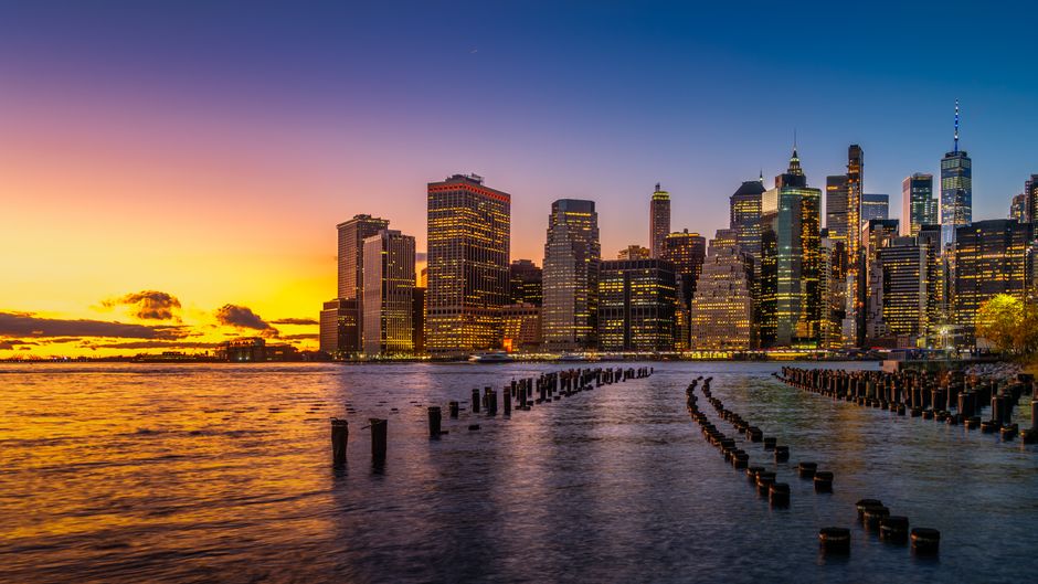 New York City skyline illuminated at sunset, viewed across the river with financial district towers visible.
