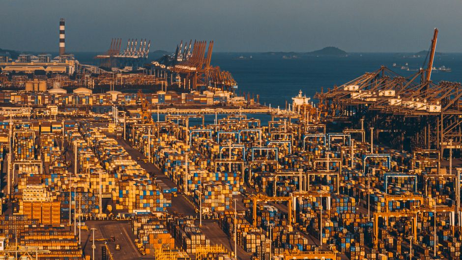 Rows of cargo shipping containers stacked at a busy port terminal with gantry cranes