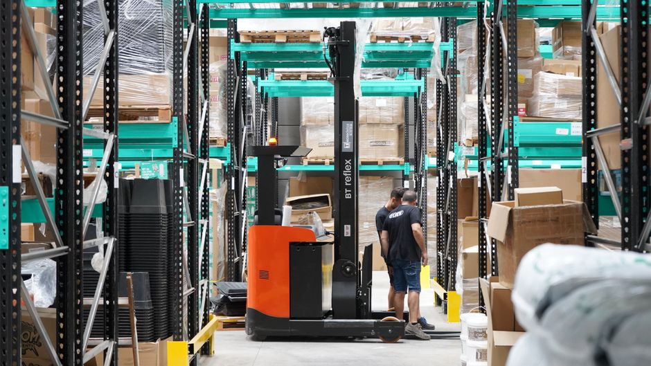 Warehouse scene featuring workers and a forklift navigating logistics aisles, representing Amazon's fulfillment and cloud infrastructure scale.