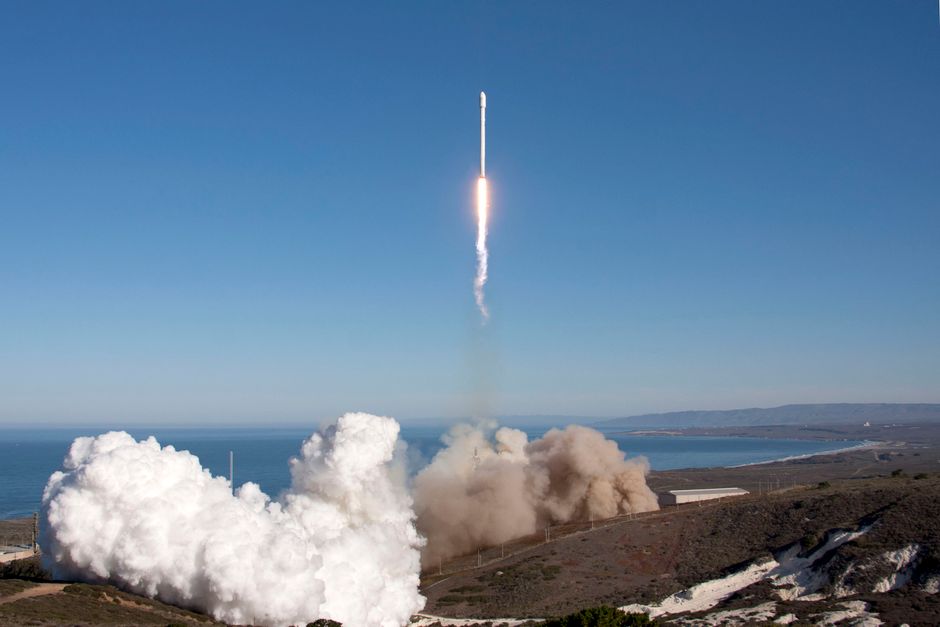 A rocket ascending on a column of exhaust against a clear sky, viewed from a distance over water